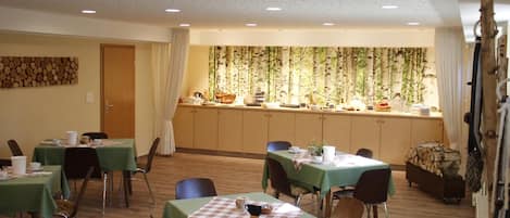 A dining area with tables set for meals, a kitchen area with a buffet, and a forest-themed wall.