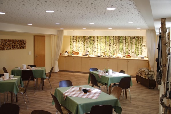 A dining area with tables set for meals, a kitchen area with a buffet, and a forest-themed wall.