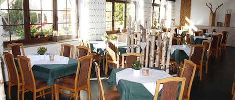 A dining area with multiple tables and chairs, large windows with curtains, and a tiled floor.
