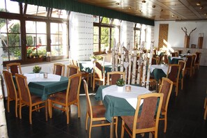 A dining area with multiple tables and chairs, large windows with curtains, and a tiled floor.