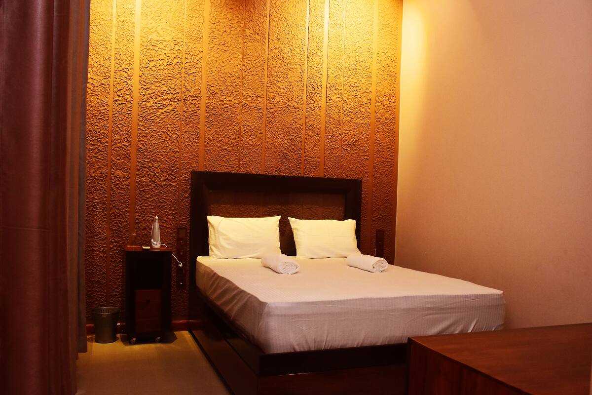 Una cama bien hecha con sábanas y almohadas blancas, una mesita de noche con lámpara y una pared texturizada detrás de la cama.