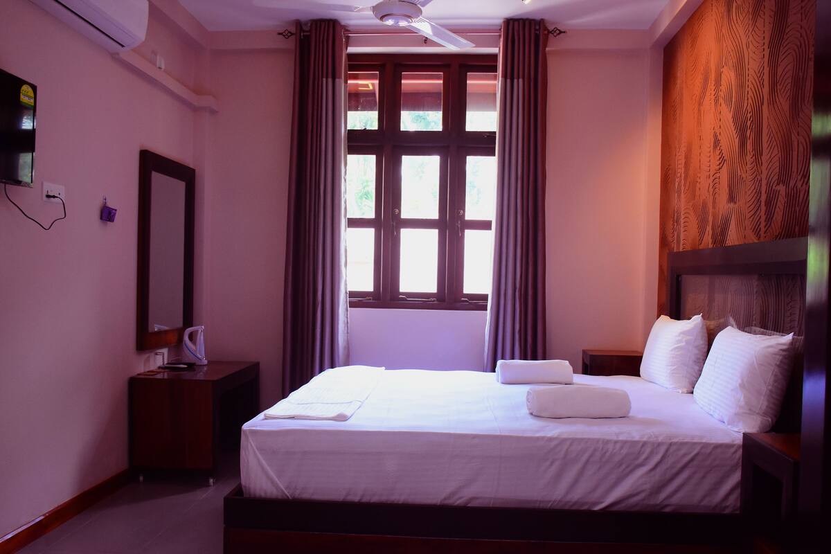 Habitación de hotel con cama, mesita de noche, espejo, ventana con cortinas y televisor de pared.