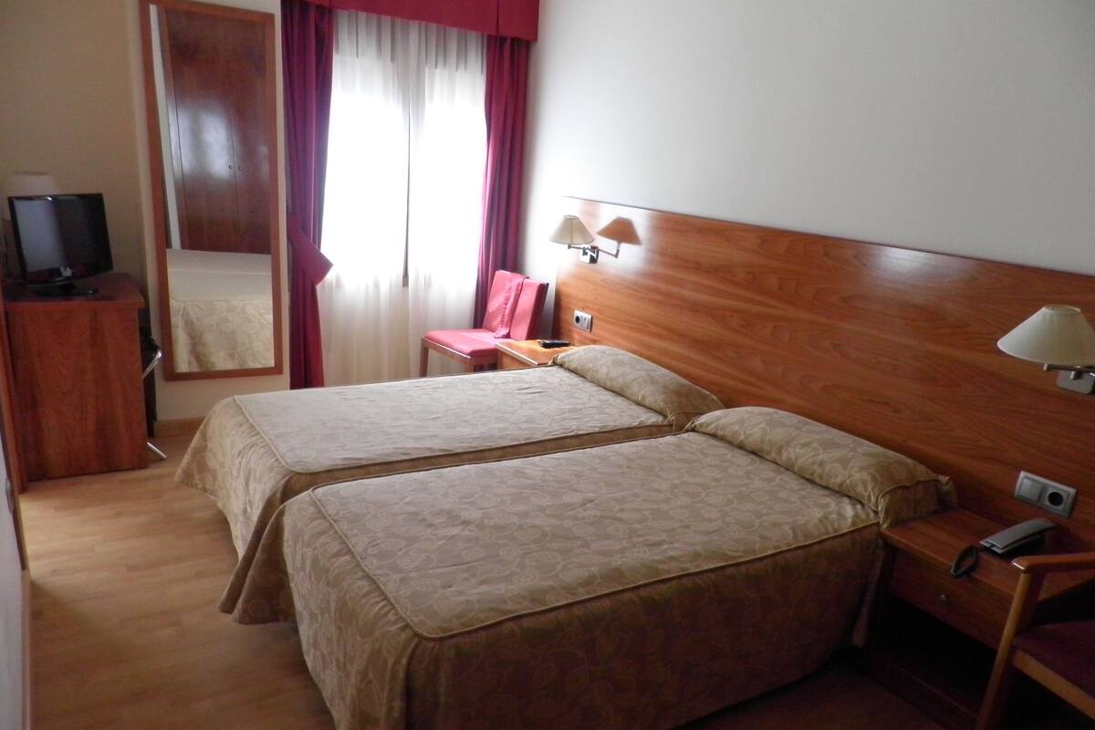 Una habitación de hotel con dos camas, un cabecero de madera, un escritorio con lámpara, una silla, un televisor sobre un mueble bajo y un espejo.