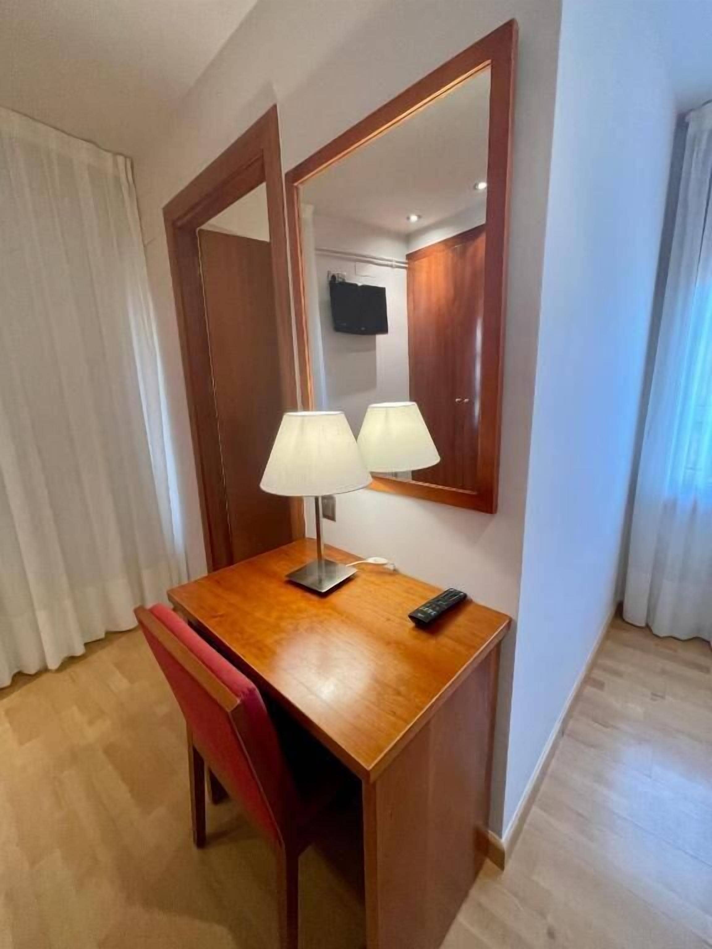 A hotel room with a wooden desk, two lamps, a red chair, a mirror, and a television on the wall.