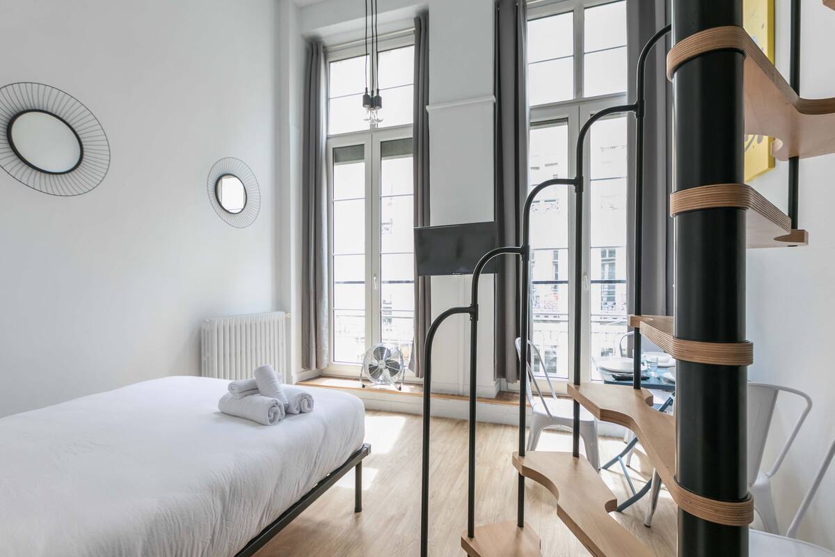 A modern hotel room with a bed, a window with a view, a radiator, and a fan.