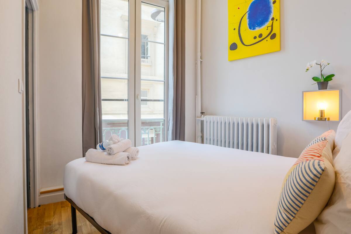 A hotel room with a bed, a window with curtains, a radiator, a wall-mounted light, and a small plant.