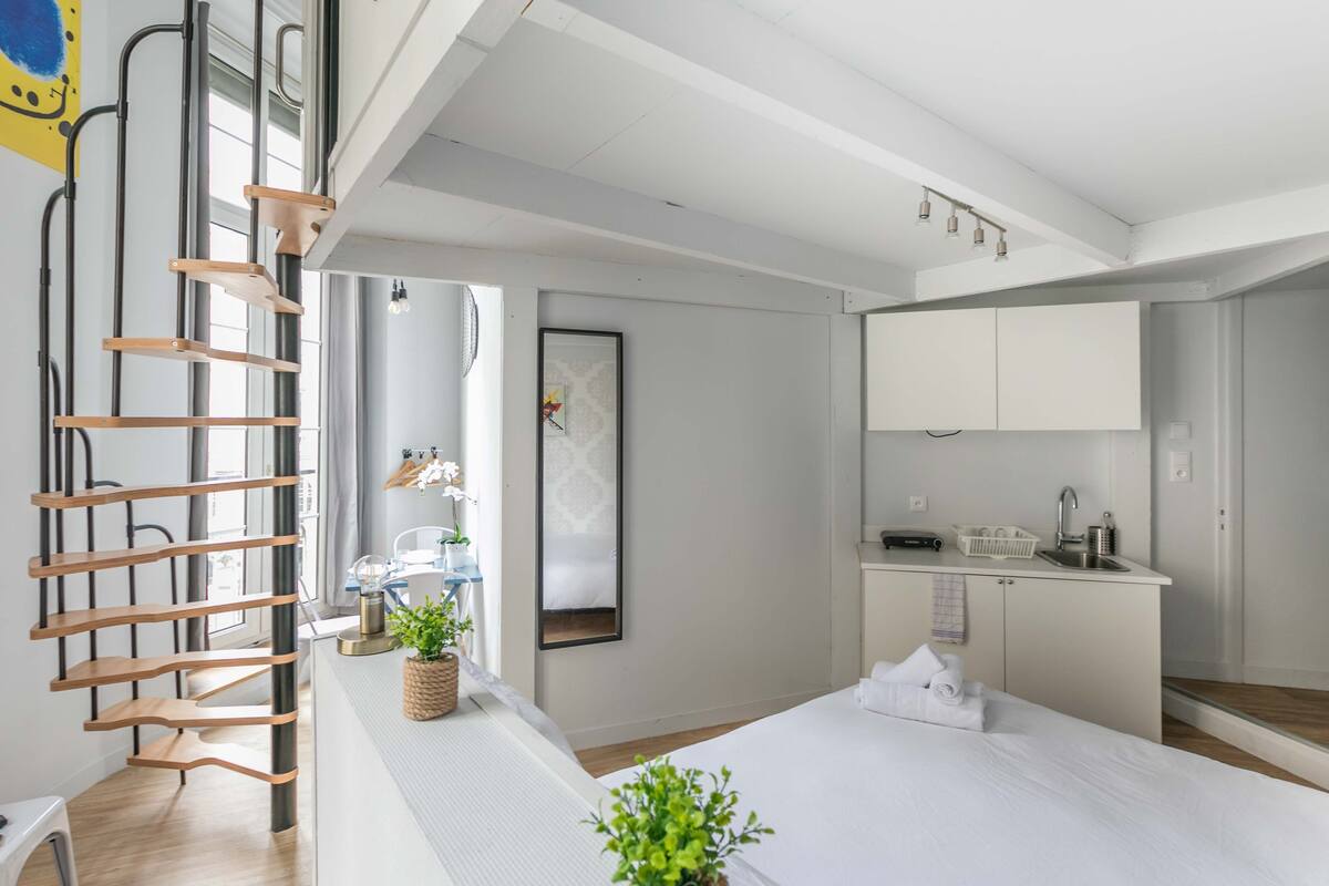 A modern hotel room with a wooden ladder, a bed, a small kitchen area, and a large window.