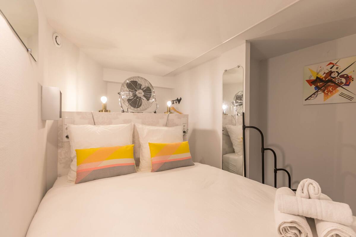 A hotel room with a bed, two pillows, a standing fan, a towel, a mirror, and a wall art piece.