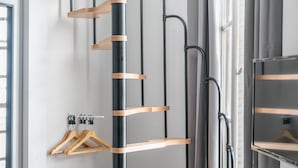 Una habitación con una estructura metálica negra que contiene perchas de madera y estantes, una ventana con persianas y un espejo en la pared.
