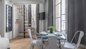 Una habitación moderna con una mesa de comedor, sillas blancas, una pequeña planta en maceta y un televisor en la pared.