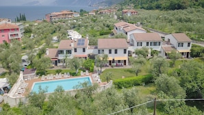 Uma área residencial com piscina, cercada por espreguiçadeiras, casas com telhados vermelhos e vista para o mar e montanhas ao fundo.
