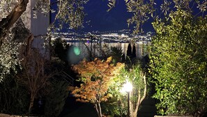 Vista noturna de um horizonte urbano com edifícios iluminados e um jardim bem iluminado, com árvores e um caminho.