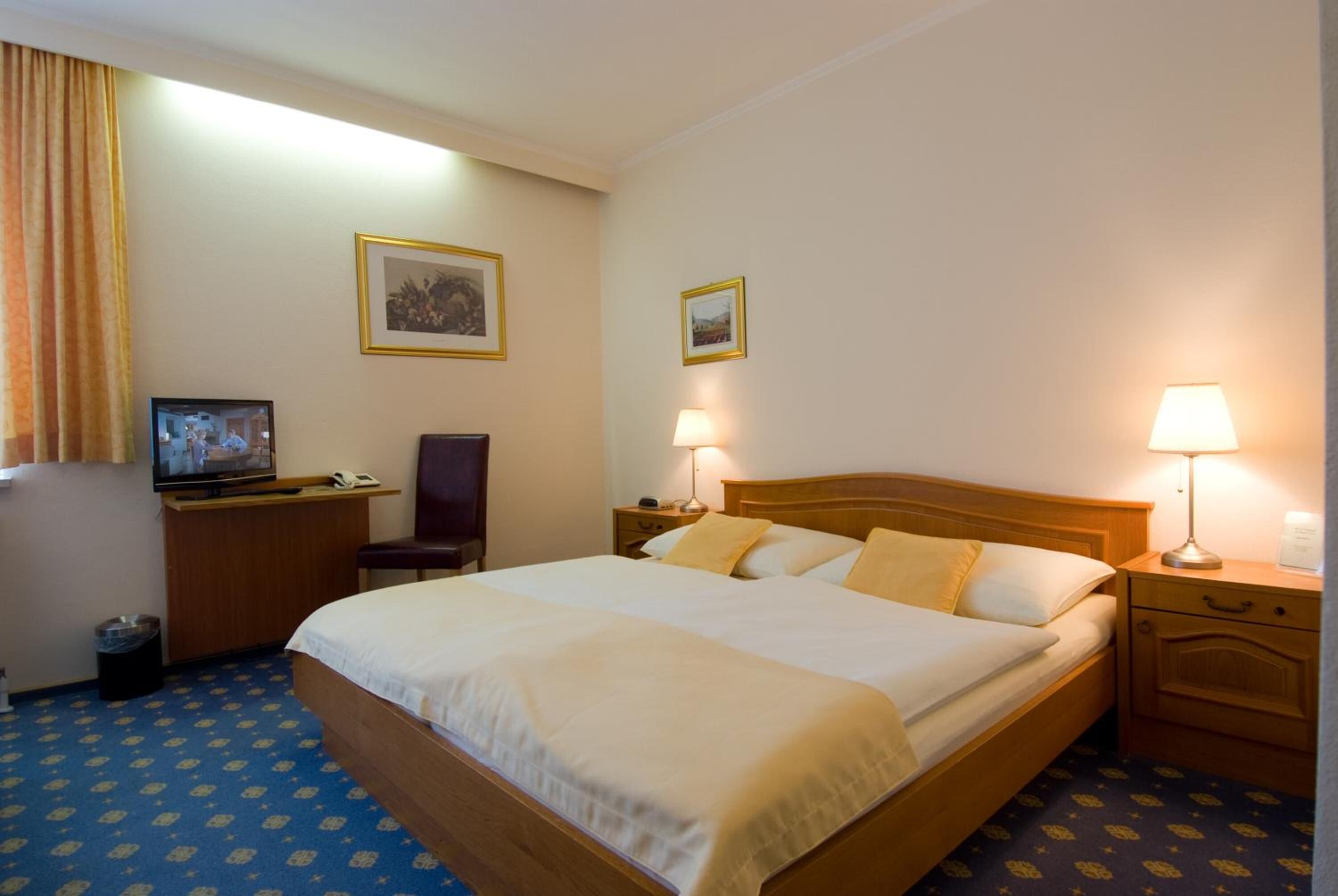 Ein Hotelzimmer mit einem großen Bett, einem Schreibtisch mit Fernseher, zwei Nachttischlampen und zwei gerahmten Bildern an der Wand.