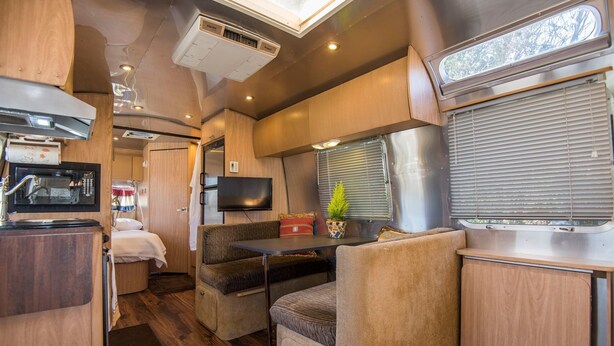 Ein kompaktes Wohnmobil-Interieur mit Küchenzeile, Essecke inklusive Tisch und Stühlen, Fernseher und Fenster mit Jalousien.