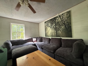 Ein Wohnzimmer mit einem Ecksofa, einem Holztisch, einem Deckenventilator und einem großen Fenster mit Blick auf Bäume.