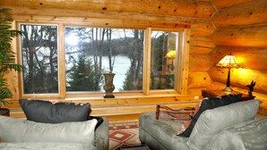 A cozy cabin interior with a large window overlooking a forest and a lake. There are two grey sofas with cushions, a small table with a lamp, and a decorative vase. - Fully furnished log home in one of northern Wisconsin's last wilderness areas (Mercer)
