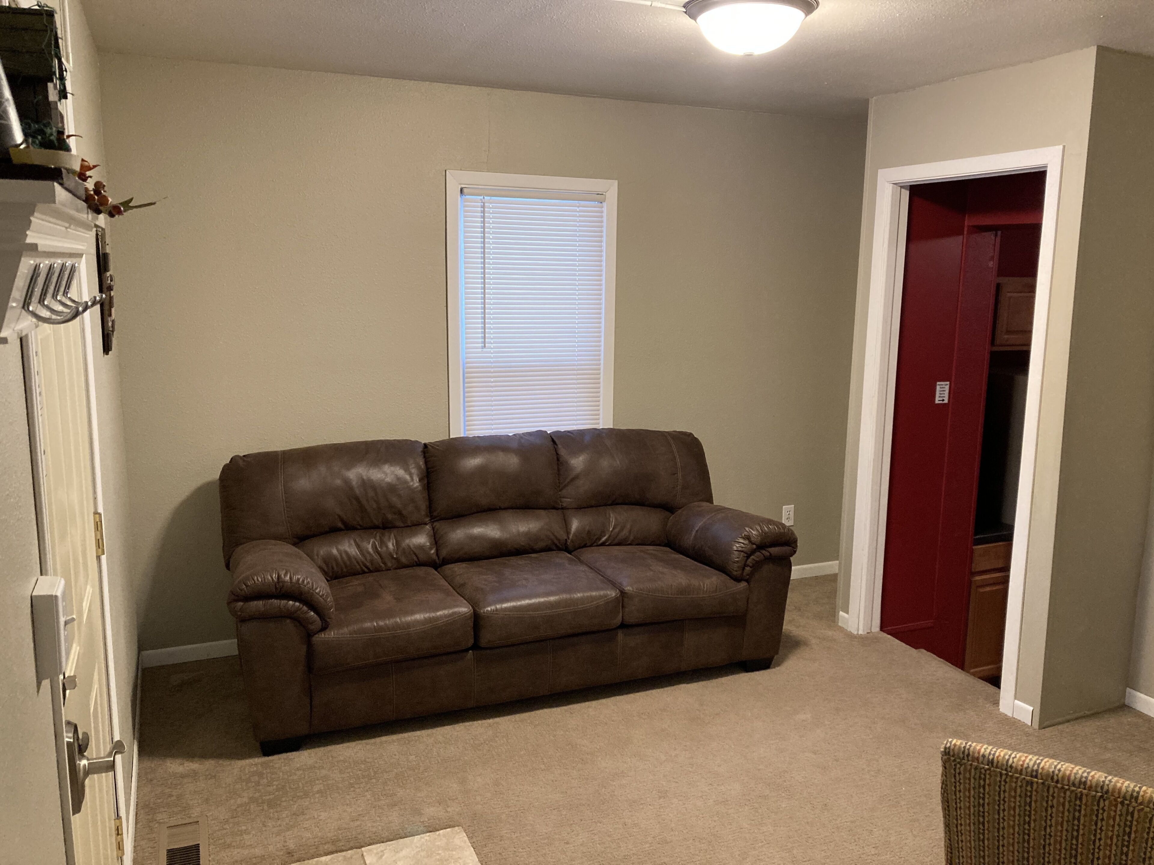 Ein Wohnzimmer mit braunem Ledersofa, beige gefärbten Wänden, einem Fenster mit Jalousien und einer roten Tür.