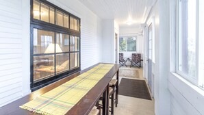 Een lange houten tafel met een geruit tafelkleed, aan weerszijden van ramen en een deur, leidend naar een terras.