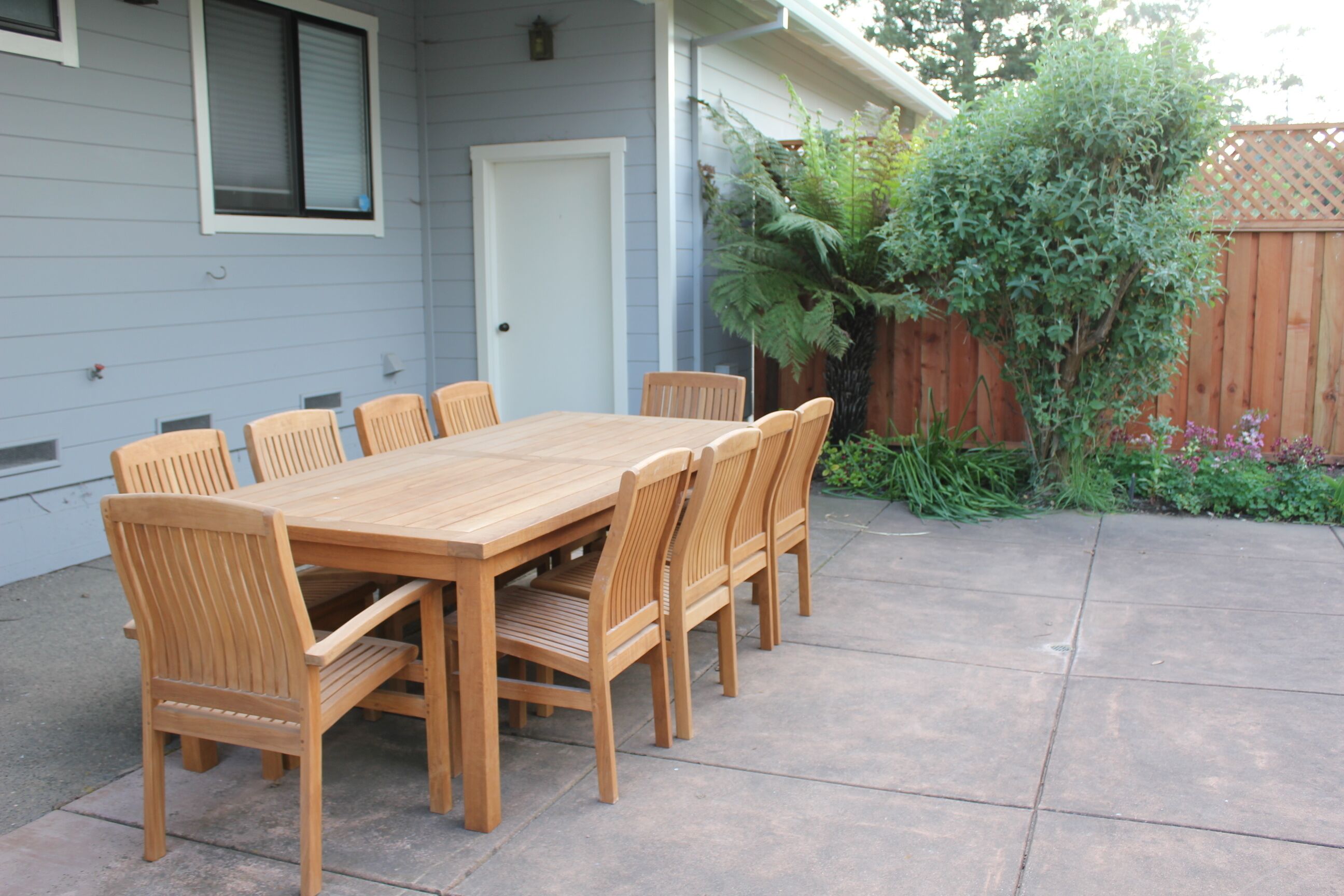 Une table à manger en bois, accompagnée de six chaises, est placée sur une terrasse en béton attenante à une maison, elle-même entourée d’une clôture en bois et de verdure.