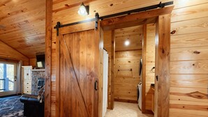 En inredning med träpanel, en skjutdörr i ladugårdsstil som leder till ett tvättrum, en eldstad i sten och en utsikt genom en glasdörr.