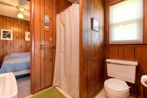 Quarto com painéis de madeira, cama, cortina de chuveiro, vaso sanitário e janela com persianas.
