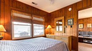 A room with wooden paneling, a large bed with a quilted cover, a dresser with a mirror, and a sofa in the background. - 401 Dog-Friendly - minutes to Beach Access & YMCA (Nags Head)