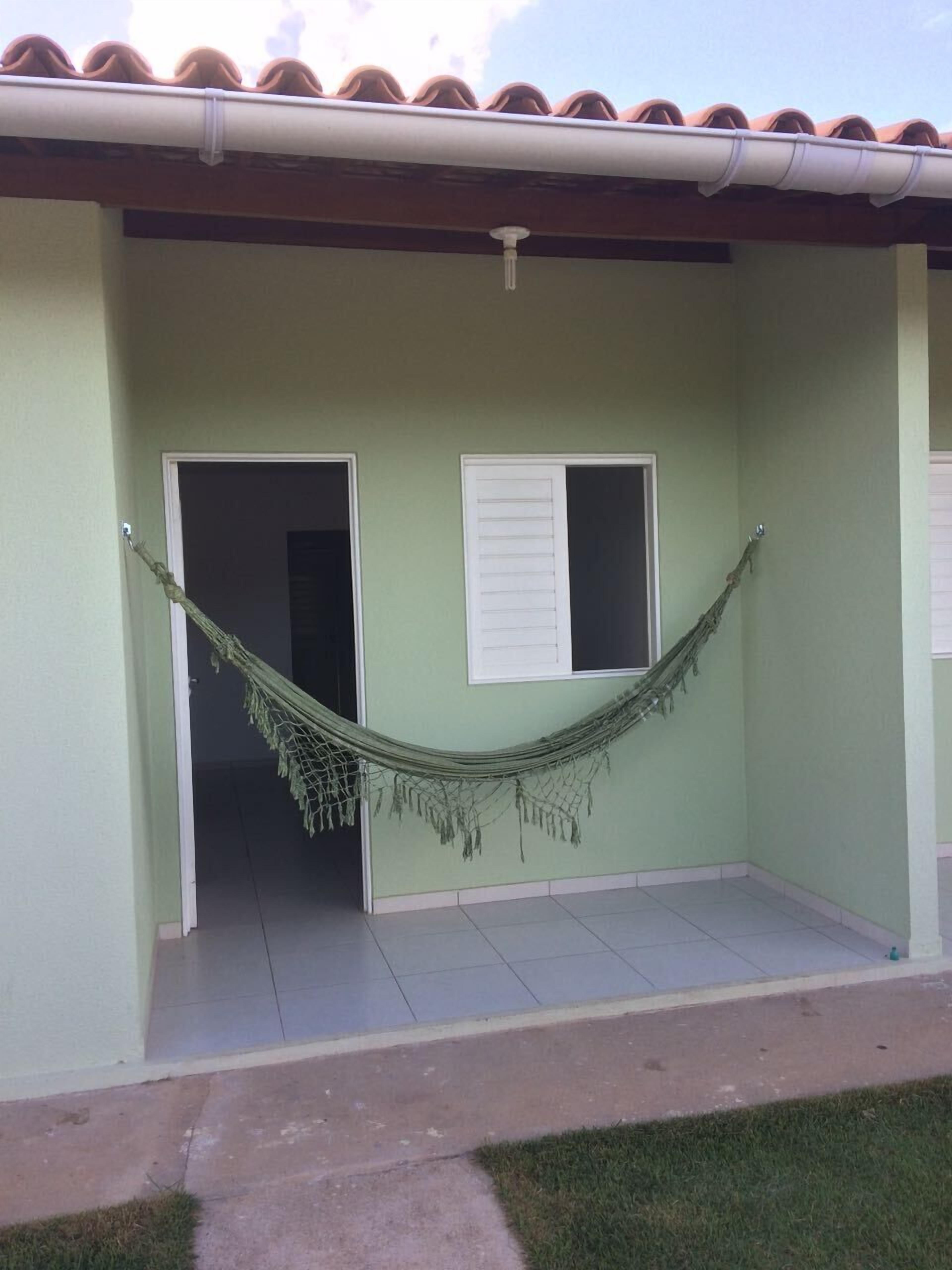 Un edificio con fachada verde clara, un porche con baldosas y una hamaca tendida entre dos puertas.