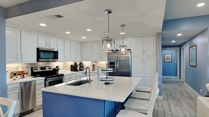 A modern kitchen with white cabinets, stainless steel appliances, a blue accent wall, and a central island with bar stools. - Stunning Ocean View Retreat w/ Spacious Balcony & Pool Access (Tybee Island)
