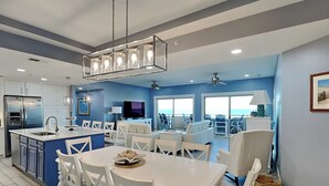 A modern kitchen with white cabinetry, a central island with seating, and a dining area with white chairs. - Stunning Ocean View Retreat w/ Spacious Balcony & Pool Access (Tybee Island)
