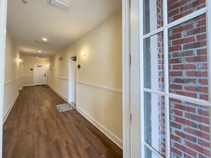 A hallway with wooden flooring, white walls, and a brick wall with windows.