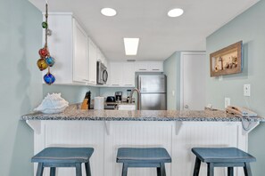 Una cocina con armarios blancos, un refrigerador de acero inoxidable, un microondas y una encimera de granito. Hay tres taburetes azules y un adorno colgante.