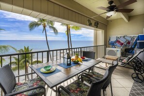 A balcony with a view of the ocean, wicker furniture, a table with a fruit bowl, and a ceiling fan. - Oceanfront Penthouse - AC, Crashing Waves, Spectacular Sunsets, Surfers, Turtles (Kailua-Kona)