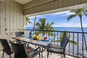 A balcony with a table set for two, overlooking a beach with palm trees and the ocean. - Oceanfront Penthouse - AC, Crashing Waves, Spectacular Sunsets, Surfers, Turtles (Kailua-Kona)