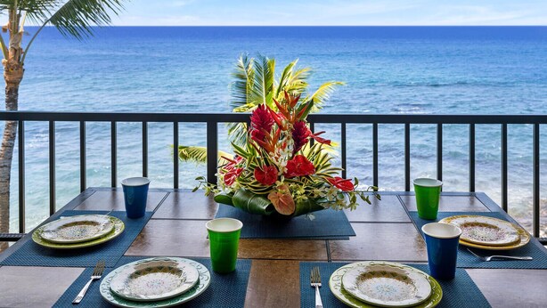 Mesa para dos personas, con platos, cubiertos y vasos, con vista a una playa, el océano y palmeras.