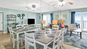 A dining area with a glass partition, white chairs, a wooden table, ceiling fans, and a view of the ocean.
