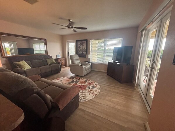 A spacious living room with a large sofa, a coffee table, and a television. There is a ceiling fan, a chair, and a window with blinds. - Stunning Key West Style Waterfront Home with Exceptional Views! (Tampa)