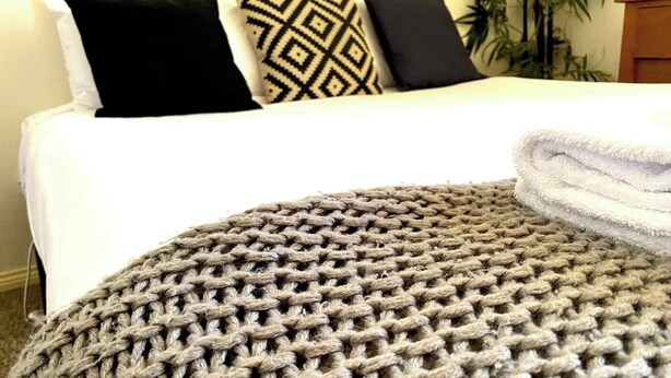 A bed with a white bedspread, two black pillows, and a patterned pillow. A grey knitted throw is on the bed. - Dora Hall | Very Central (Orange)