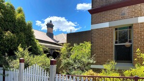 A brick building with a white picket fence, a window, and a chimney. - Dora Hall | Very Central (Orange)