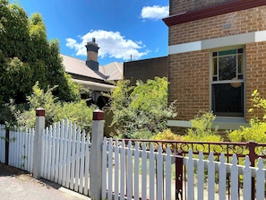 A brick building with a white picket fence, a window, and a chimney. - Dora Hall | Very Central (Orange)