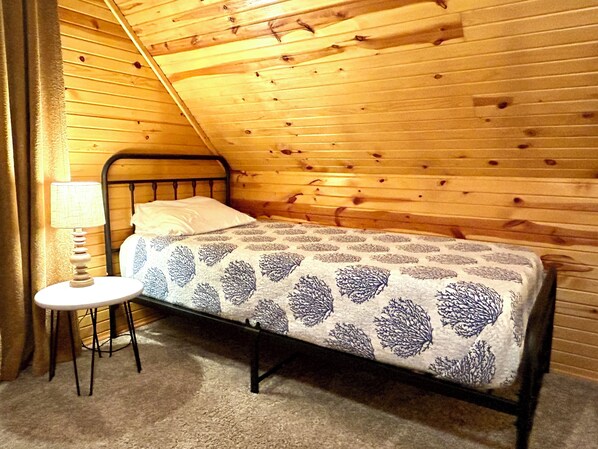 A single bed with a patterned bedspread in a room with wooden walls and a sloped ceiling. - Privacy and Luxury await! Fire Pit Patio, Large Outdoor Deck, and Heated Pool. (Mears)