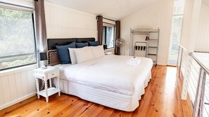 A bedroom with a large bed, a bedside table, a lamp, a window with curtains, a fan, and a drying rack.