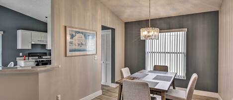 A dining area with a wooden table and chairs, a chandelier, and a window with blinds.