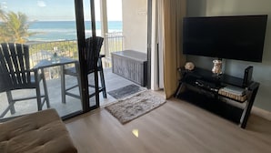 Een woonkamer met een grote televisie, een bank, een balkon met stoelen en uitzicht op de oceaan.