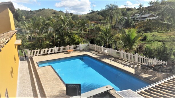 A rectangular swimming pool with a white fence and a bench, surrounded by palm trees and a hilly landscape. - Leisure and Rest for any day (Guararema)