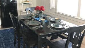 Una mesa de comedor con platos, tazas y cubiertos, un refrigerador negro, y sillas en una habitación con ventana y adornos en las paredes.