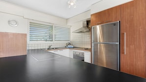 Une cuisine moderne dotée d’un réfrigérateur en acier inoxydable, de meubles en bois, d’un plan de travail noir et d’une fenêtre avec des stores.
