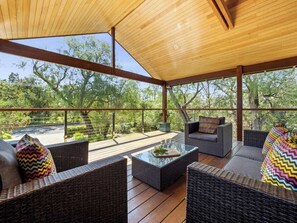Un patio al aire libre con mobiliario de mimbre, una mesa con superficie de vidrio y vistas a árboles y al cielo.