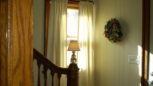 A staircase with a wooden handrail, a wall-mounted lamp, a window with white curtains, and a floral wreath.