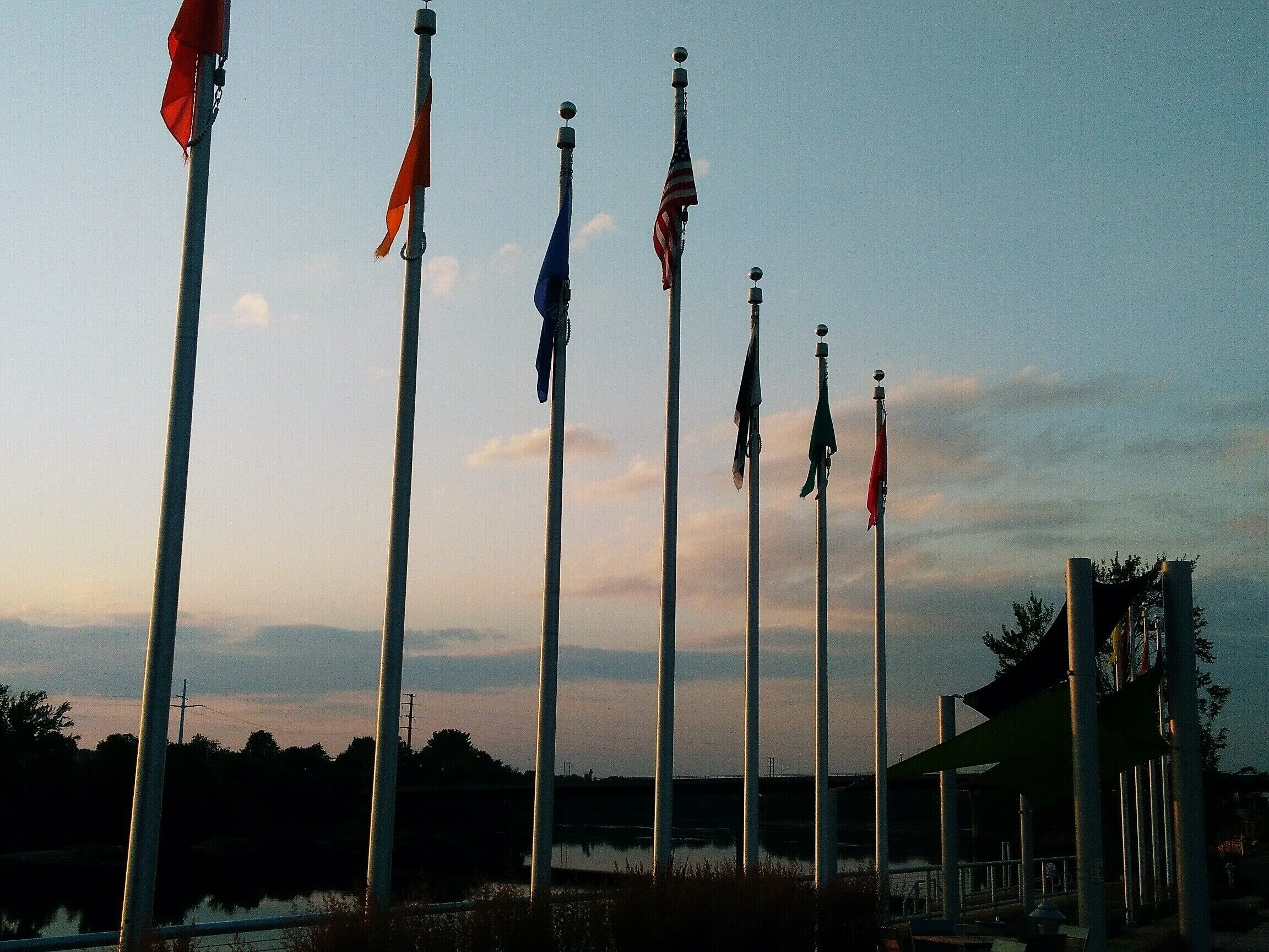 A row of flagpoles with flags, a body of water, and a building in the background.