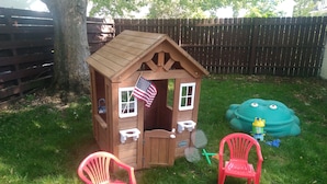 A wooden playhouse with a flag, two red plastic chairs, and a green inflatable toy.